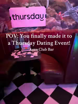 The image shows the interior of a bar or club. A sign with the word "thursday" in a pink rectangle hangs from the ceiling. Below the painted brick wall, there is a booth. The foreground shows a black and white checkered floor, suggesting a retro or vintage theme. A table with scattered cards sits in front of the booth.