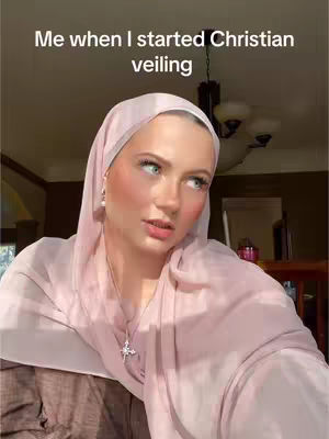 A woman wearing a light pink hijab looks up and to the side with a somewhat surprised expression. She is wearing a silver necklace with a cross pendant and pale pink blush. The background is out of focus but appears to be a well-lit room.
