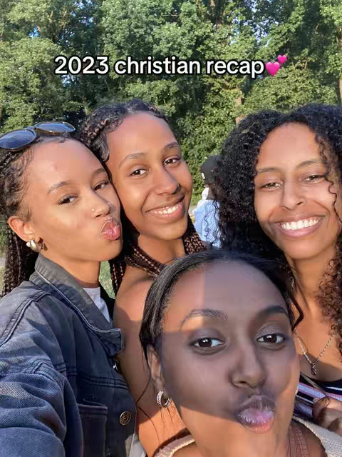 A selfie-style image featuring four young women posing closely together. The women are fair-skinned, with varying hairstyles, and are smiling or making playful faces at the camera. The background shows blurred green foliage, suggesting an outdoor setting. The overall composition is bright and captures a sense of camaraderie.
