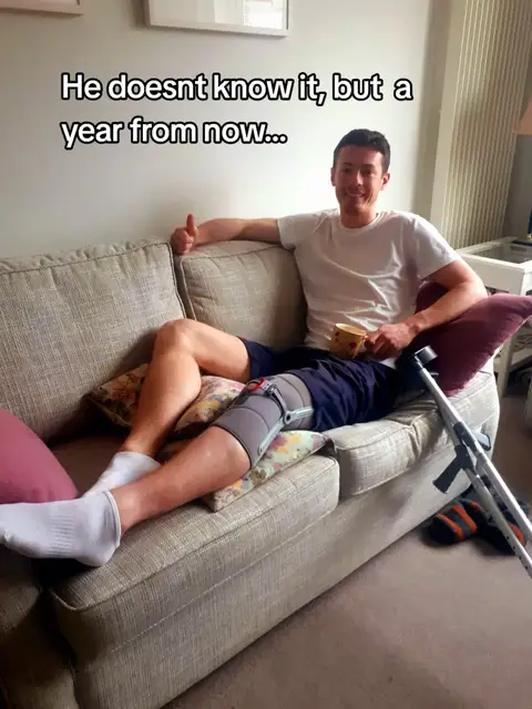 A man is reclining on a gray couch, his leg propped up on pillows. He has a knee brace on one leg and is giving a thumbs up. Text overlayed. Crutches are to the side.