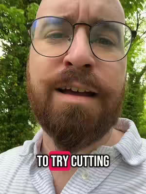 A close-up shot of a man with glasses and a trimmed beard, facing the camera. The background is blurred green foliage.