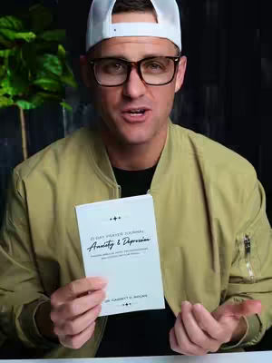A man wearing glasses and a white baseball cap holds a small, white book in his hands. He is smiling and looking towards the camera. Behind him is a blurry background with green foliage.
