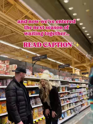 A couple stands in a grocery store aisle. The woman is laughing and leaning forward, dressed in a white hat and black coat. Her partner, wearing a black cap and jacket, seems to be responding. The background shows store shelves with products. The lighting is bright.