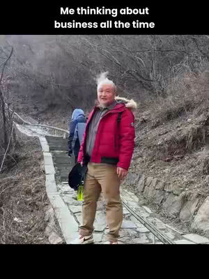 An older gentleman stands on a stone pathway with a winding staircase behind him, set in a natural environment. The man is wearing a red jacket, tan pants, and sneakers. His hair stands up in a unique pattern, with the text overlayed at the top.
