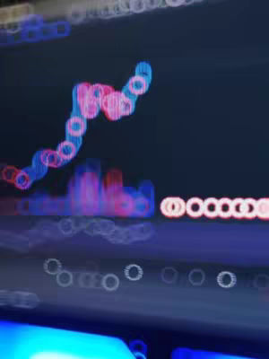 An out-of-focus image of what appears to be a digital display, possibly a stock chart, with blurry circular highlights in shades of blue and pink. The background is dark, suggesting a low-light or night setting. The composition is abstract, using light and color to create an impression of data visualization without clear details.