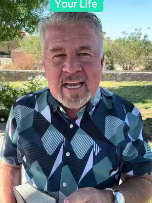 A middle-aged man with gray hair and a beard is centered in the frame, facing the camera with a slight smile. He wears a patterned, short-sleeved shirt. Above his head, a rectangular shape with the words "Your Life" is in bright green. The background suggests an outdoor setting, with trees and a bright sky visible.