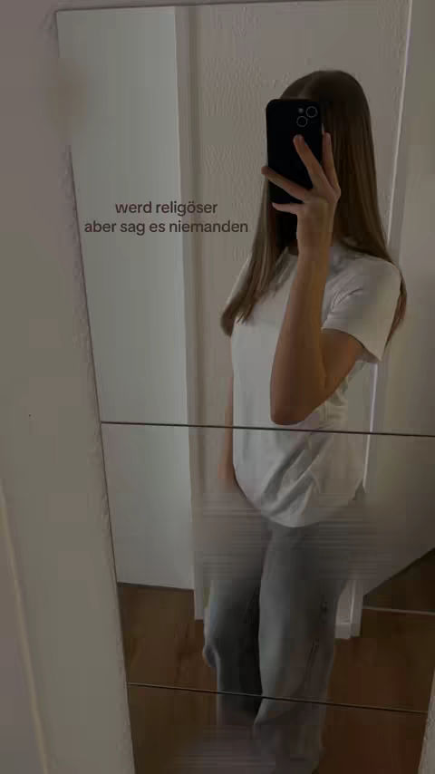 A person is taking a selfie in a mirror, obscuring the face with a phone. The image is mostly grayscale, showcasing a white t-shirt and gray pants. Text is overlaid on the image in white.