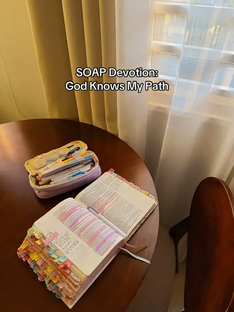 A top-down shot featuring an open Bible on a wooden table, its pages marked with colorful sticky notes and highlighting, next to a pencil case with pens and markers. The scene is illuminated by natural light from a nearby window.