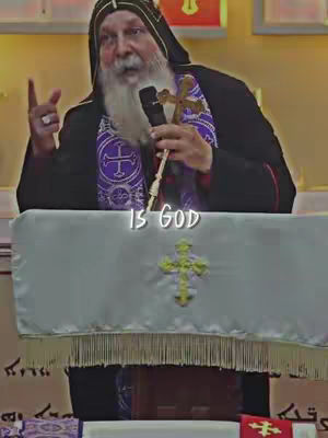 An older man with a long white beard is speaking into a microphone. He's wearing a black robe with a purple collar and a black head covering. Behind him, a simple golden cross is attached to a microphone. The overlay displays 'IS GOD' in a casual white font.