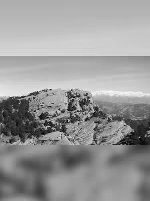 A black and white photograph of a rocky mountain peak with some trees. The sky is a light gray, the mountain dark gray, and in the background are more mountains and a few clouds. The focus is on the rock formation.