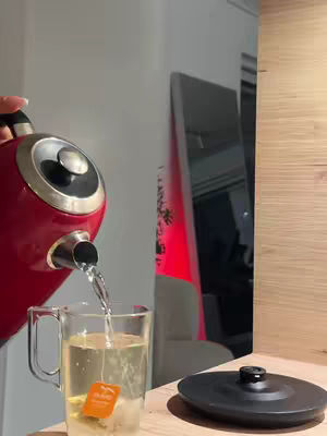 A close-up shot shows a red kettle pouring hot water into a clear glass mug with a tea bag. The cover shows a tea being prepared, with part of a wooden surface and a clear glass in view. In the background, a mirror reflects some ambient light and nearby furniture.