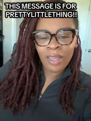 A woman with long, reddish-brown locs, wearing large black-framed glasses and a black top, looks directly at the camera with a neutral expression. The background is a softly lit, nondescript interior space.