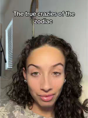 A selfie-style shot featuring a woman with curly dark hair and subtle makeup, positioned against a neutral-toned background. Text overlayed over the top-left corner reads 'The true crazies of the zodiac'. The woman is looking at the camera with a neutral expression.