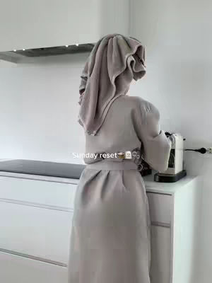 A person is standing with their back to the camera, wearing a grey robe and a towel wrapped around their head. They are facing a white cabinet with a coffee machine on top. The background is a minimalist white room.