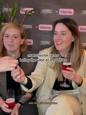 Two women are shown smiling while interacting with a microphone and holding glasses of red liquid, likely wine or a cocktail, in front of a backdrop with logos. The woman on the left is holding the microphone, and the woman on the right is holding a glass. Both women have light skin, and the one on the left has blond hair and the one on the right has brown hair. The lighting appears to be from overhead.