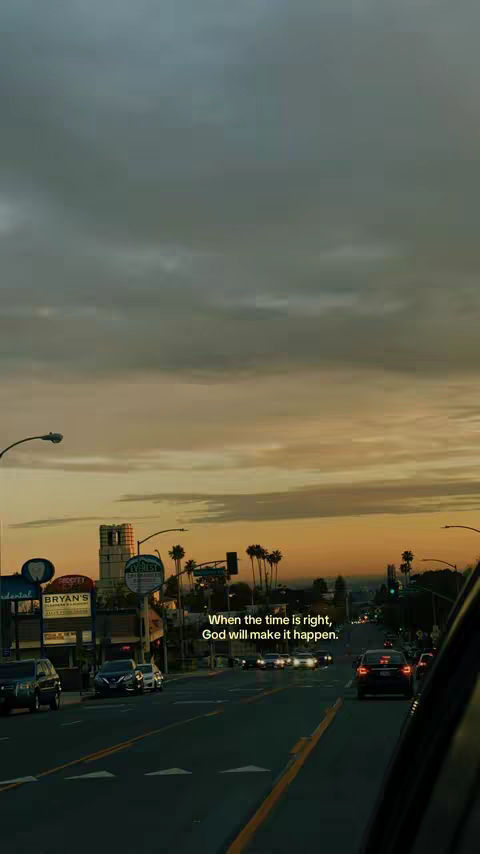 A shot from inside a car looking down a city street at sunset. The sky is a mix of gray and golden hues. Buildings and streetlights line the road.