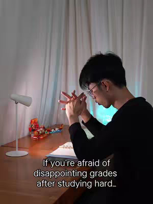 The video depicts a young man seated at a wooden desk, hands clasped together, with a focused expression. He is wearing glasses and a black sweater. A desk lamp and a colorful Lego set are visible. Text overlayed in the lower part reads, "If you're afraid of disappointing grades after studying hard..."