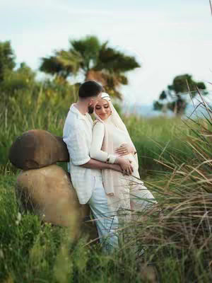 A romantic outdoor shot of a couple embracing. The woman, wearing a white hijab and dress, is pregnant and has her hands on her belly. The man, in a white shirt, embraces her. They are in a field with green grass, tall weeds, and a blurred background of trees.