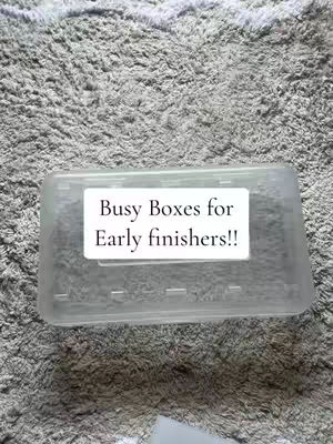 A clear plastic box is presented diagonally on a textured, gray surface that resembles a carpet or woven material. A white rectangular label is affixed to the box, centered and displaying the text 'Busy Boxes for Early finishers!!' in a simple, black font.