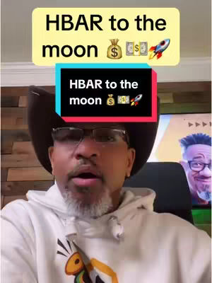 A selfie-style video cover image featuring a man wearing glasses, a cowboy hat, and a white hoodie. Above his head are two text overlays. The top one is a yellow rectangle with the text 'HBAR to the moon' and various emoji. The second overlay is a black rectangle with the same text and emojis. The man has a look of excitement on his face. In the background, there's a room with a TV screen, where a face is seen. The overall composition is bright and centered on the man's face.