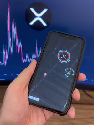 A hand holding a black cell phone, which displays a map-like interface with an XRP logo on the screen. In the background, there's a blurred chart and the XRP logo is visible.