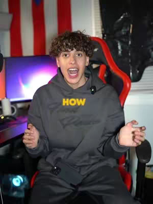 A young man with curly hair is in the foreground, sitting in a red and black gaming chair. He is wearing a gray hoodie with the word "HOW" in yellow across the front. His mouth is open in an excited expression; his arms are spread out to the sides. Behind him is a monitor and the flag of Puerto Rico is on the wall.