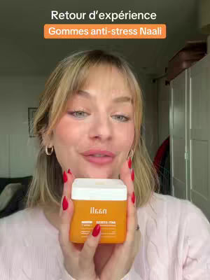 A woman with blonde hair and red painted fingernails holds a white and orange product container close to the camera. Text overlays appear above the woman's head in an orange box. The background is a softly lit interior with a cabinet and glimpses of a framed picture.