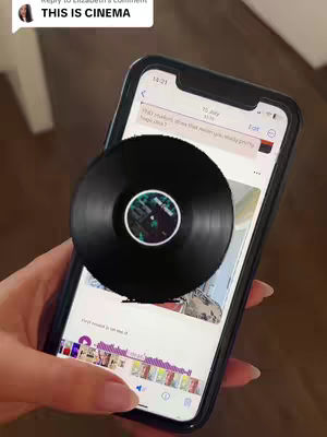 A hand holding a smartphone is the central element of the image. The phone display shows an app interface along with a record overlay. The record is black with a central label. The background is blurred, suggesting a warm indoor setting. The overall tone is visually appealing.