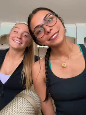 A duo of young women smile directly at the camera. One on the right wears glasses and has a braid. The other on the left has long blonde hair and a black top. The background is a neutral-toned interior setting.