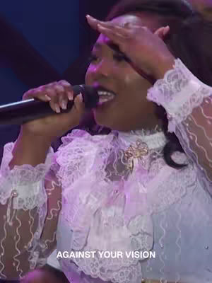 A woman with dark hair is centered, singing into a microphone with her right hand raised to her forehead. She wears a white, frilly blouse with lace details. The background is a blurred mix of dark and light colors, and the overlaid text reads, 'AGAINST YOUR VISION'.