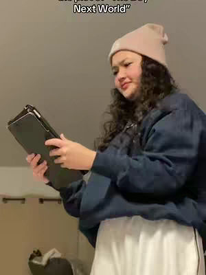 A woman with curly hair wearing a pink beanie and a dark blue jacket is holding a dark-colored tablet, looking slightly to the side with a subtle expression. The background is a simple, neutral-toned wall.