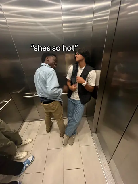 Two young men stand in an elevator, facing each other and smiling. The walls of the elevator are metallic and reflective. A text overlay at the top says "shes so hot".