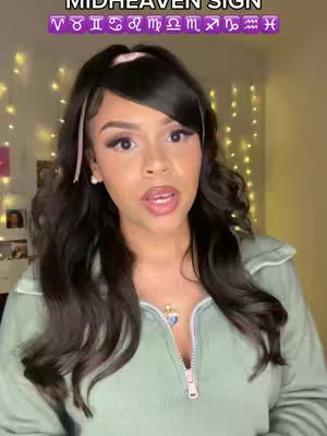 A woman with long, dark hair and makeup is facing the camera and talking. She is wearing a light green zip-up, a necklace with a blue heart pendant, and small, white earrings. Above her head is a purple banner with zodiac signs in white text.
