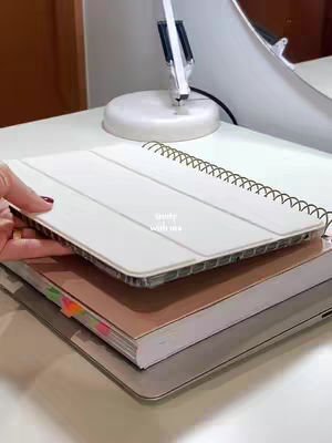 A close-up shot of a white desk with a stacked notebooks and a desk lamp. The camera focuses on a hand holding open a notebook, with the text 'study with me' on the notebook itself. The overall composition is clean with neutral color tones and a focus on the notebook and desk setting.