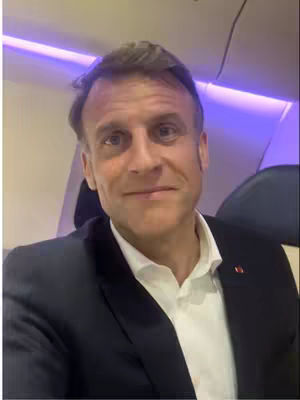 A selfie-style shot inside an airplane cabin. The central figure is a man with short brown hair, wearing a navy suit jacket over a white shirt. The background shows the interior of the plane with muted purple lighting overhead.