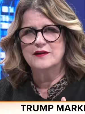 A medium shot shows a woman with glasses, light-colored hair, and red lipstick. A panel displays the text 'TRUMP MARK'. The background is blue.