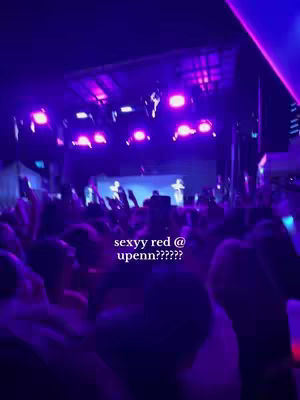 A low-angle shot from within a crowd, looking towards a brightly lit stage. In the foreground, a sea of blurred faces and raised hands. The stage is illuminated with purple stage lights and spotlights. Text overlayed on the screen includes the name "sexyy red" and the location "upenn." The overall atmosphere conveys excitement and energy.