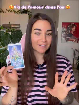A woman with long brown hair and a pink and black striped shirt holds up a tarot card, with her hands open as she talks. The background includes a blurry indoor setting with green plants and a framed painting on the wall. Text overlay with hearts in the upper area.