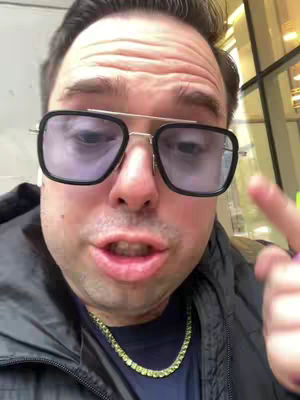 A close-up shot of a man wearing large, square-framed glasses, with a black jacket and a gold chain. He is outdoors, with a blurred background, and appears to be speaking directly to the camera. The image is well-lit, focusing on his face and upper body.