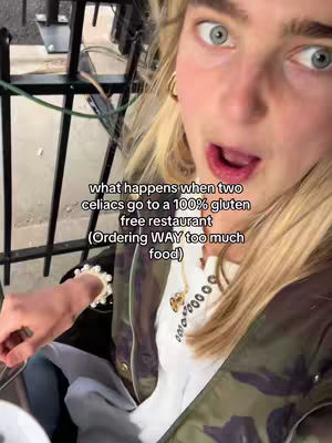 A close-up shot of a woman with a surprised expression, mouth open, showcasing her eye and facial features. She is wearing a white top, a layered jacket, and a necklace. The background includes metal bars and other indistinct elements.