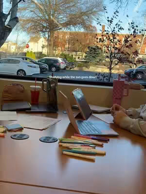 The image captures a brightly lit study scene set inside a room with a large window offering an outdoor view. The primary focus is on a wooden table filled with studying materials: a laptop, papers, pens, markers, a glass of water with a straw and a red liquid, and two cellphones. The window provides natural light, and the outside view includes cars parked along the street.