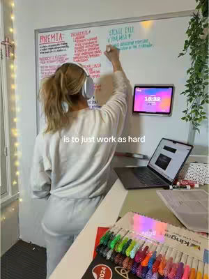 A woman wearing headphones is standing with her back to the camera, writing on a whiteboard. A tablet displays a colorful interface, and a laptop is open on a table. Text overlay reads 'is to just work as hard'.
