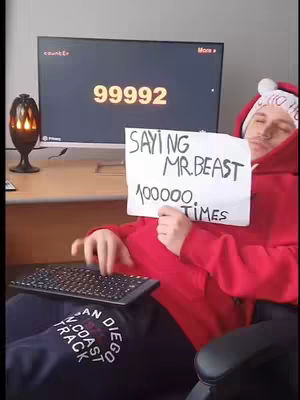 A man in a red hoodie and a Santa hat is sitting, holding a sign that says 'Saying Mr. Beast 100000  Times'. A computer monitor to his left displays the number 99992. A keyboard is in his lap.