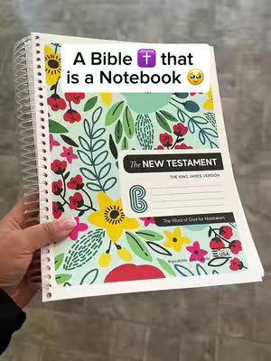 A close-up shot of a spiral-bound notebook that is open, displaying a colorful floral design. The notebook appears to be a Bible, with the text 'The New Testament' visible. The open notebook is held by a hand, with a solid gray surface as the background.