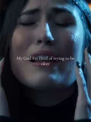 A close-up of a woman's face showing extreme emotion. Her eyes are closed, tears are visible, and her hands are near her face. Overlay text in a cursive font reads, "My God I'm tired of trying to be okay."