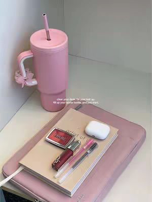 The image showcases a collection of aesthetically arranged items on a pink surface, likely a desk or table. Dominating the frame is a large pink insulated tumbler with a straw, positioned on the upper left. Beside it, are notebooks, makeup, and accessories, all in a matching soft pink palette. The composition is clean and organized, with a minimalist appeal, possibly aimed at promoting a study or work-related aesthetic. The lighting conditions are evenly distributed, enhancing the vibrant colors and overall visual charm.
