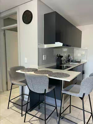 A photo showcasing a modern kitchen and dining area. A white counter extends from a dark cabinet that houses the sink and stovetop. Above, sleek black cabinets are mounted on the wall. Three gray bar stools with black legs are situated at the counter. In the background, a white door and the interior door frame are visible.
