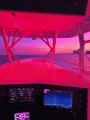A view from inside a boat, looking out at a vivid pink and purple sunset over the water. The boat's dashboard and roof structure are visible.