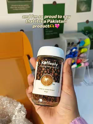 The cover image shows a hand holding a jar of coffee. The jar has a white lid and a label with the brand name "Koffeely." The label also features a coffee bean design and text. Behind the hand are items, including some papers stuck to a wall, partially visible, and a container holding pens. Overlayed text at the top of the image reads "and I'm so proud to say that it is a Pakistani product".