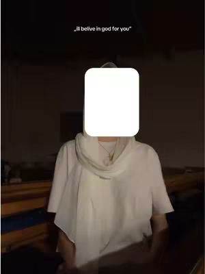 A person is standing in what appears to be a church. Their face is covered by a white square. They are wearing a white shirt and a scarf. The background is dark, the text overlay says "I'll belive in god for you".